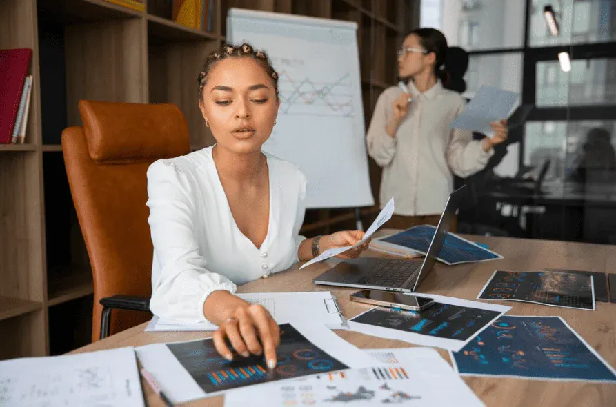 Mulher empresária analisando projetos em reunião de negócios, com colega ao fundo, em ambiente de escritório moderno com computadores e documentos.