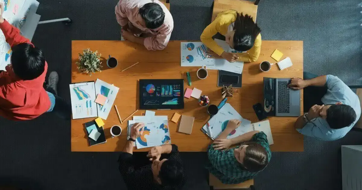 Reunião de equipe em escritório moderno com várias pessoas discutindo projetos, utilizando laptops, tablets e documentos para planejamento estratégico.