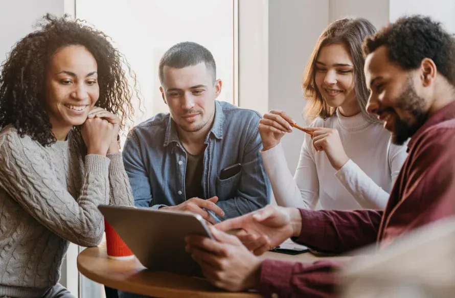 Grupo de jovens adultos sorrindo e interagindo enquanto usam um tablet em um ambiente interno acolhedor, promovendo conexões sociais e aprendizado colaborativo.