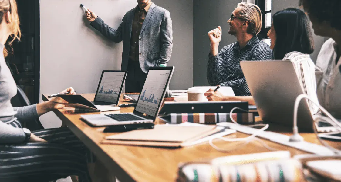 Reunião de negócios em uma sala de reunião com profissionais usando laptops, discutindo estratégias enquanto um facilitador apresenta informações na lousa branca.