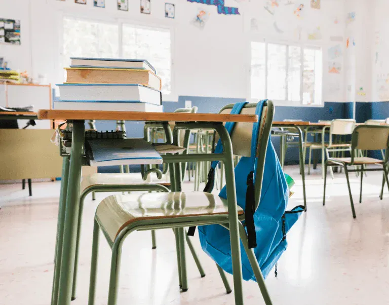 Sala de aula com mesas, cadeiras e mochila azul, ambiente escolar bem iluminado com janelas grandes e paredes decoradas com desenhos infantis.