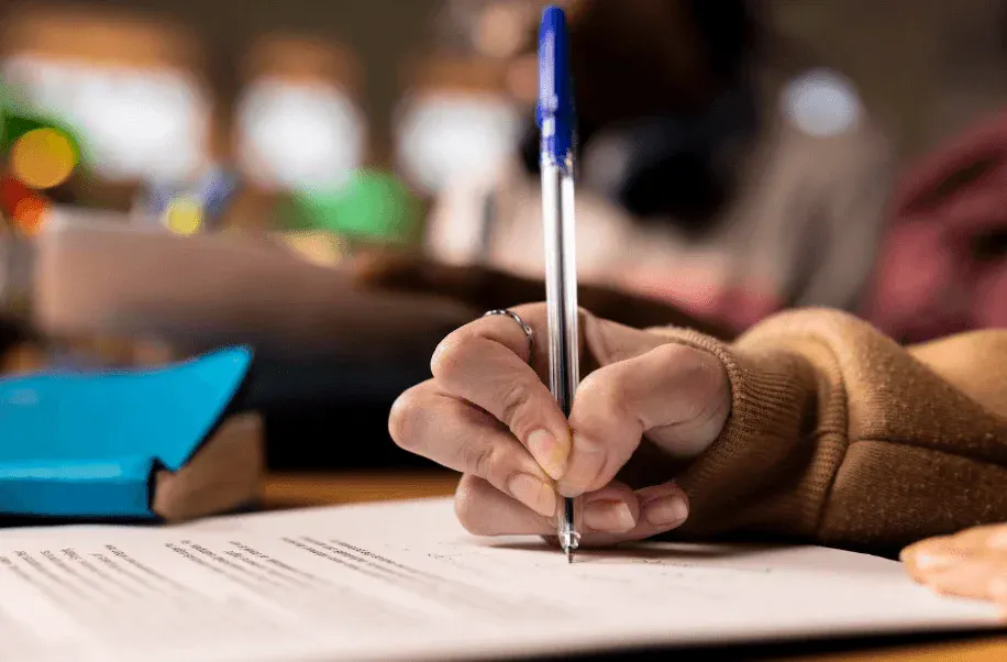 Pessoa fazendo anotações com uma caneta azul em um caderno, sentado em um ambiente de estudo ou trabalho, com foco na mão e no objeto de escrita.