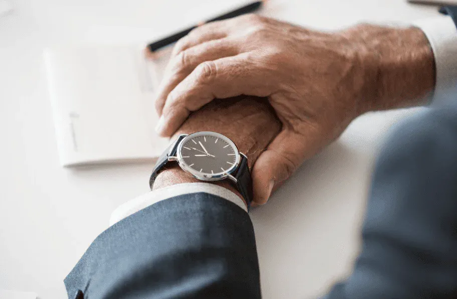 Homem de terno preto usand orelha de relógio clássico de pulso, segurando a mão de uma pessoa idosa em um ambiente de escritório.