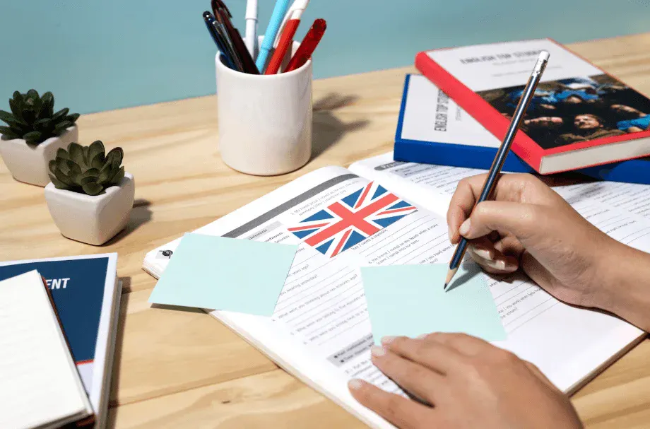 Pessoa estudando língua inglesa com materiais de estudo, incluindo livros, notas adesivas e bandeira do Reino Unido, em uma mesa de madeira.