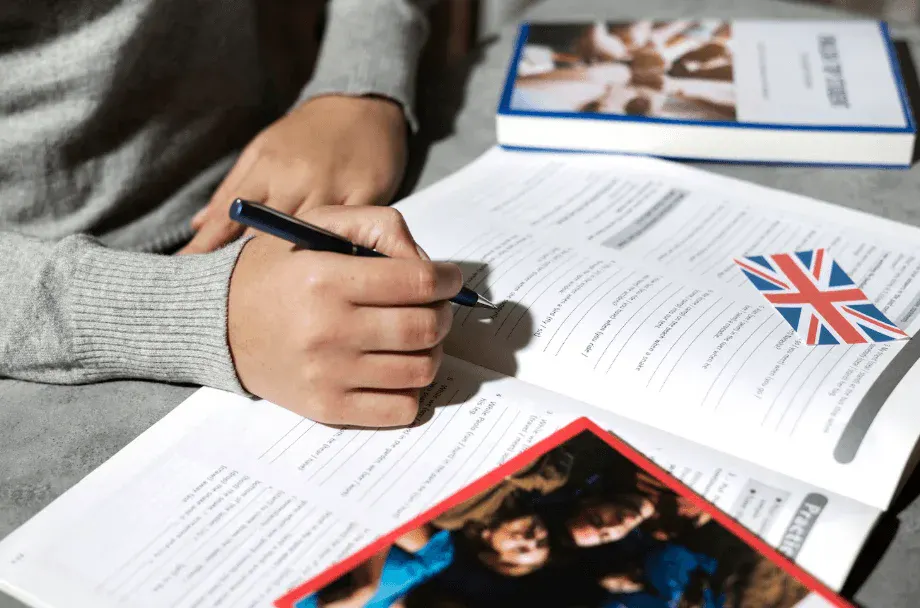 Pessoa escrevendo em uma folha de papel com uma caneta, com materiais de estudo ao redor, incluindo livros e uma bandeira do Reino Unido, retrato de estudo ou preparação de documentos.