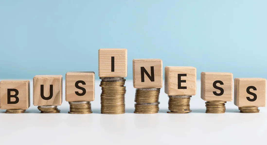 Imagem mostrando blocos de madeira com letras formando a palavra 'BUSINESS' empilhados sobre moedas, simbolizando crescimento e sucesso nos negócios.