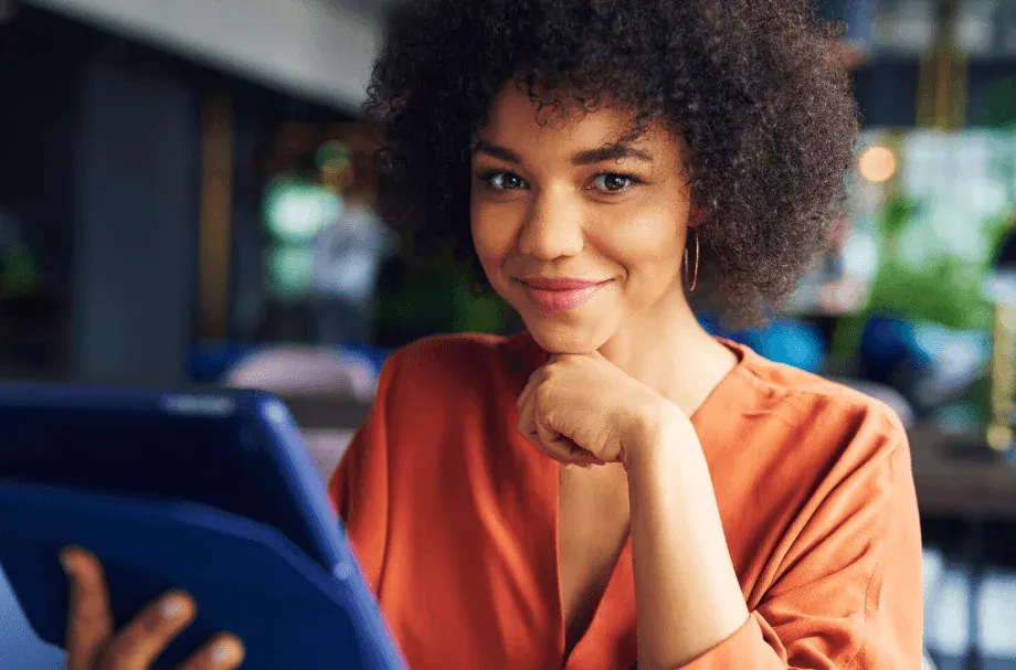 Mulher jovem sorrindo usando camiseta laranja e brincos, segurando um tablet, sentada em um ambiente aconchegante e bem iluminado.