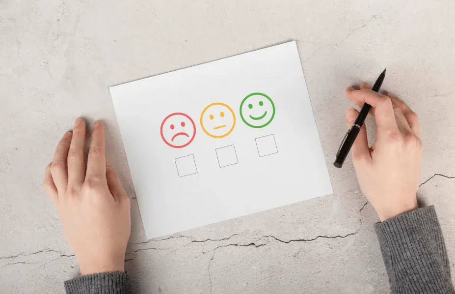 Pessoa avaliando satisfação com uma pesquisa de feedback usando emoji de rosto triste, neutro e feliz, disposta sobre uma mesa de concreto.