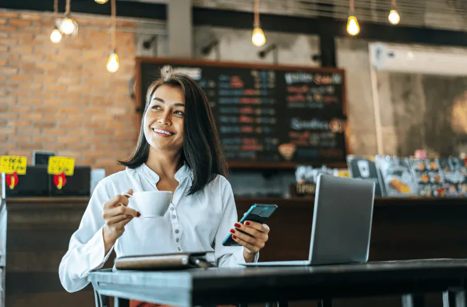 Mulher sorridente usando um celular em um café com uma xícara na mão, ambiente com iluminação quente e decoração rústica.
