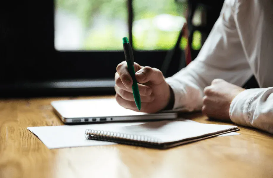 Pessoa escrevendo com caneta sobre papel, com laptop ao lado.
