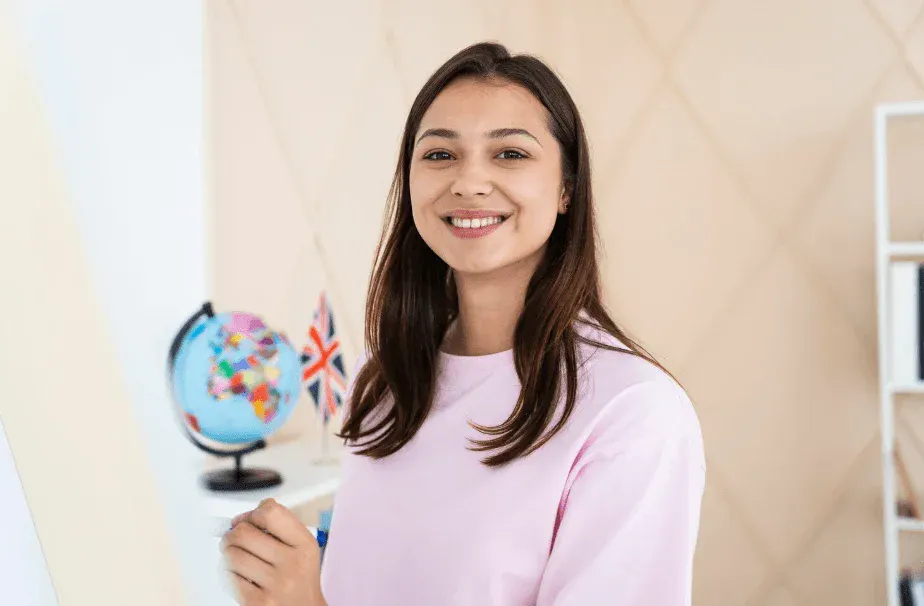 Jovem mulher sorrindo em um ambiente de sala de aula, com uma pequena bandeira do Reino Unido e um globo terrestre, representando educação e cultura internacional.