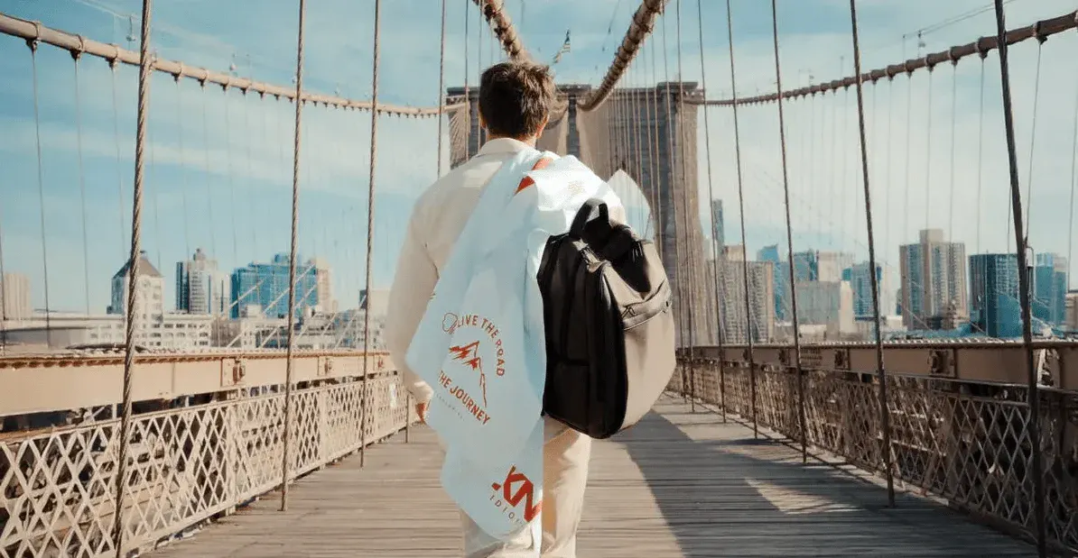 Pessoa caminhando na ponte do Brooklyn em Nova York, com mochila nas costas e cidade ao fundo, durante o dia.