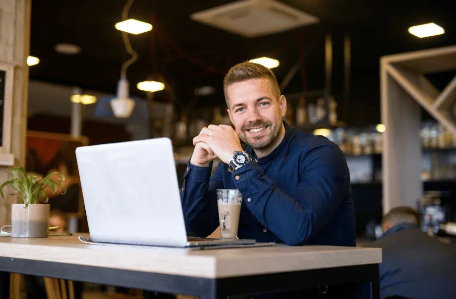 Homem sorridente sentado em uma cafeteria com laptop, bebida e ambiente aconchegante ao fundo.