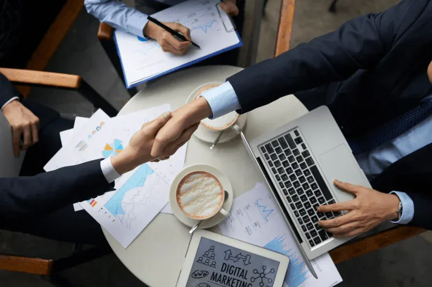 Imagem de duas pessoas apertando as mãos durante uma reunião de negócios, com gráficos, um laptop e tablets com conteúdo de marketing digital na mesa.