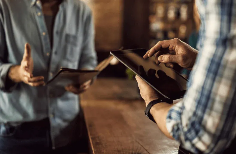 Imagem de duas pessoas usando tablets em ambiente moderno, possivelmente discutindo ou negociando, destacando o uso de tecnologia e comunicação.
