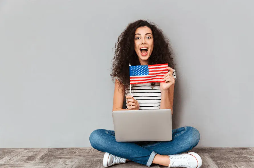 Mulher jovem sentada no chão com laptop, segurando uma bandeira dos Estados Unidos.