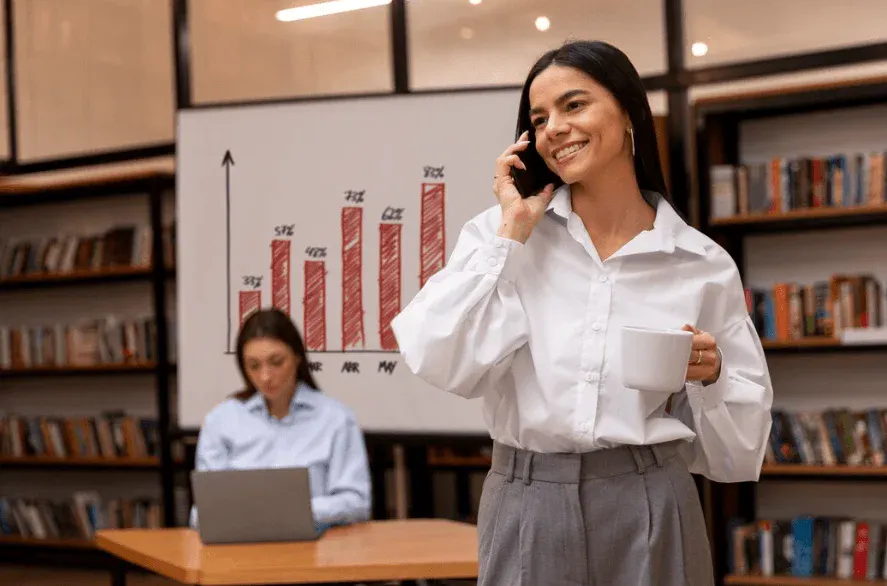 Mulher sorridente conversando ao telefone enquanto segura uma caneca, ao fundo uma mulher trabalhando no laptop em ambiente de escritório com gráficos e livros