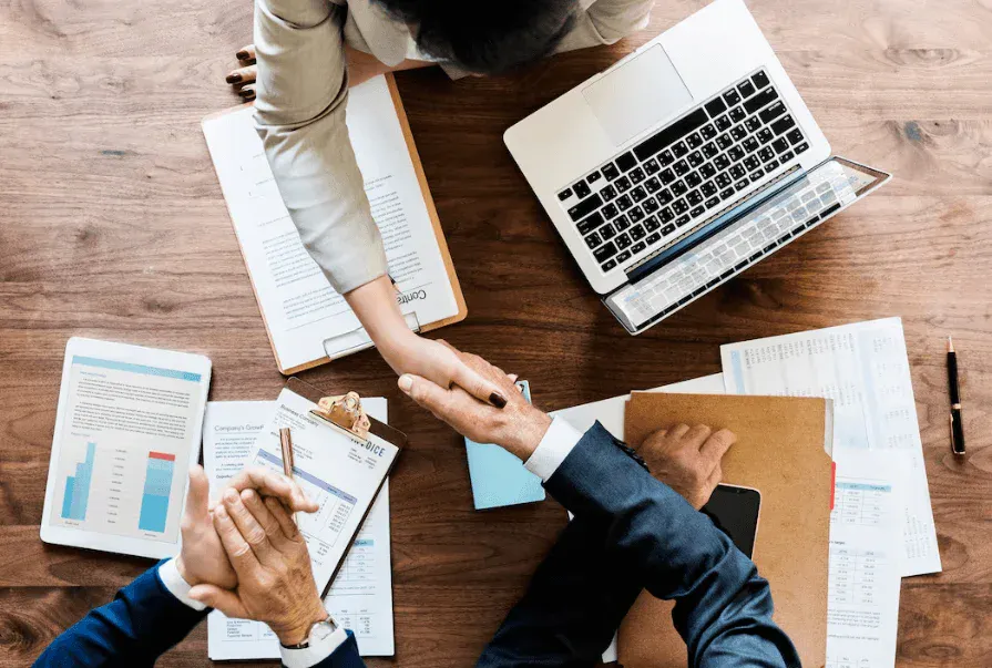 Duas pessoas apertando as mãos em negociação de negócios na mesa de escritório com documentos, laptop e smartphone, simbolizando parceria empresarial.
