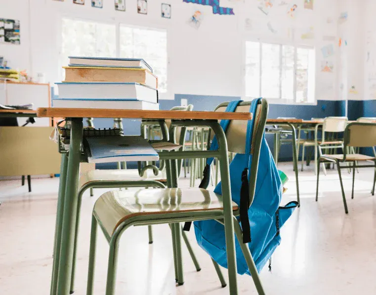 Imagem de uma sala de aula com cadeiras, mesas, livros e uma mochila azul pendurada. Ambiente escolar iluminado com janelas e paredes decoradas com desenhos e cartões.