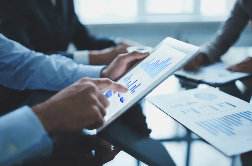 Reunião de negócios com pessoas analisando dados em uma mesa, utilizando tablets e papéis com gráficos e informações financeiras, ambiente corporativo.