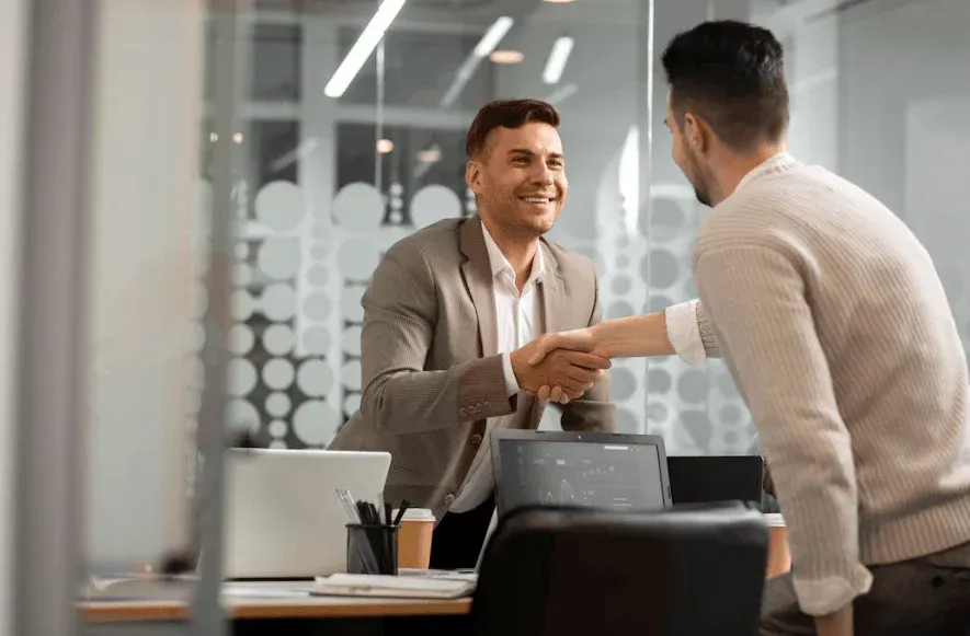 Homem de terno apertando a mão de outro homem em escritório moderno, simbolizando acordo ou parceria de negócios.
