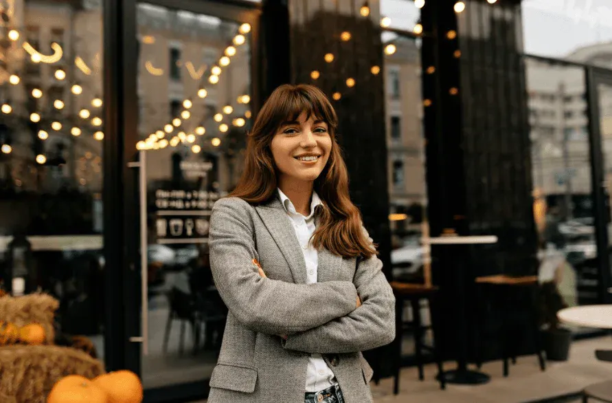 Mulher sorridente com cabelo castanho e roupas formais, posando em frente a uma loja moderna com iluminação aconchegante durante o dia.