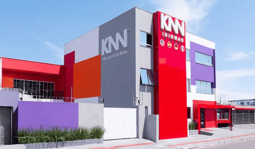Fachada do centro de idiomas KNN Idiomas, edifício moderno com cores vibrantes vermelho, roxo e cinza, fachada com o logo da marca, localizado em uma área urbana com céu azul ao fundo.