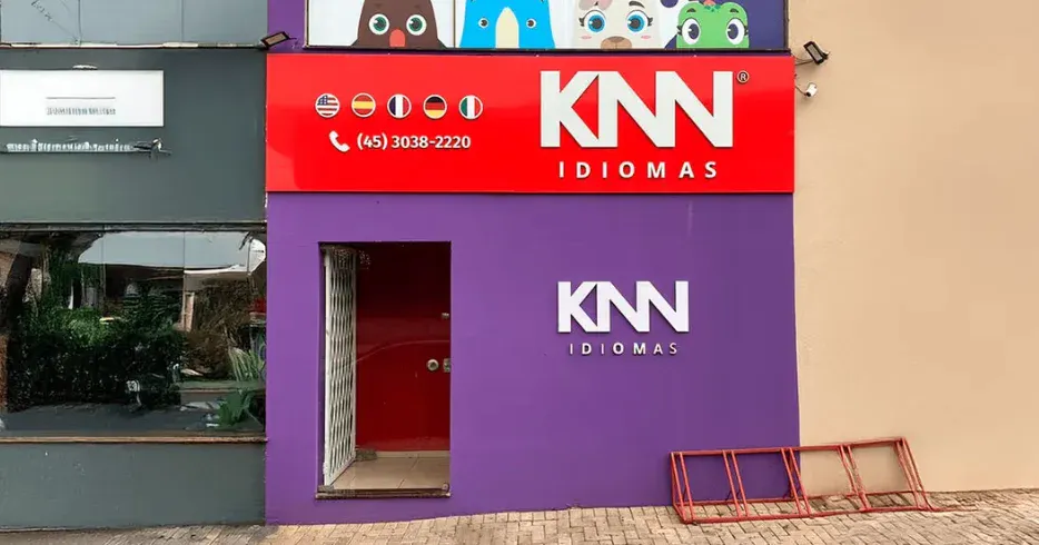 Fachada da escola de idiomas KNN Idiomas, com cores vibrantes em roxo e vermelho, e painel com ícones de bandeiras de diversos países.