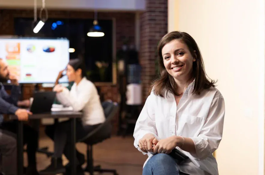 Imagem de uma jovem mulher sorridente em ambiente de escritório, com pessoas se reunindo ao fundo, representando um ambiente de trabalho colaborativo.