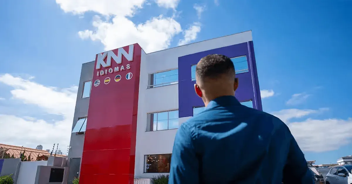 Pessoa olhando para o edifício da escola de idiomas KNW Idiomas com um céu azul ao fundo, promovendo ensino de idiomas para diferentes públicos.