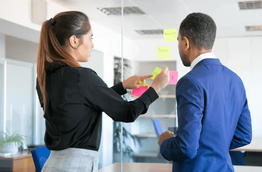 Dupla de profissionais discutindo estratégias de marketing digital em ambiente de escritório, usando post-its coloridos para organizar ideias.