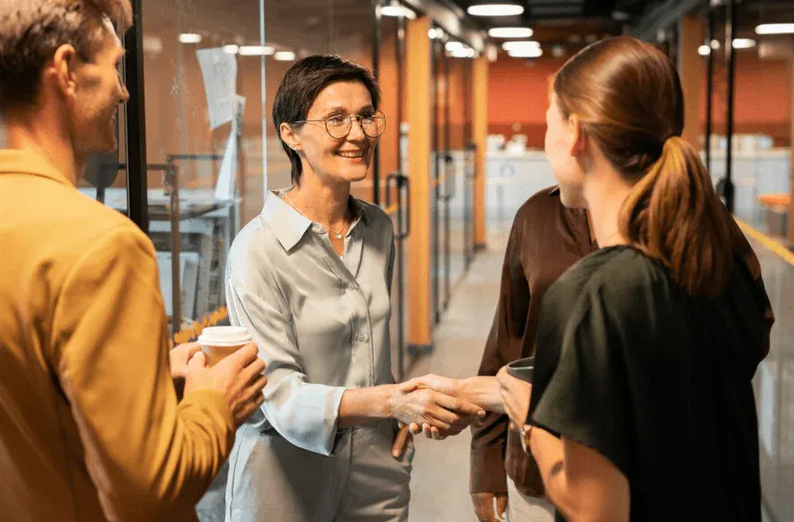 Mulher empresária sorridente cumprimentando outras pessoas em ambiente de escritório moderno, com colegas de trabalho ao redor, promovendo networking.
