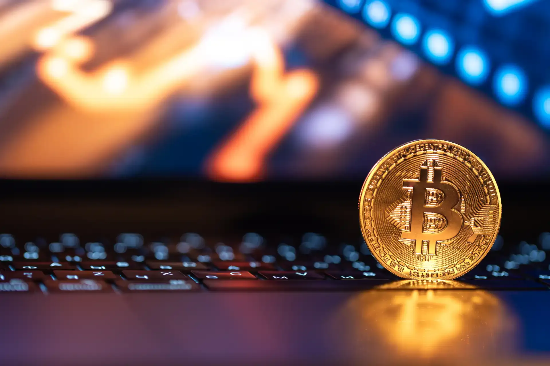 Imagem de uma moeda de Bitcoin dourada sobre o teclado de um laptop, com uma tela de computador ao fundo com gráficos e dados relacionados a criptomoedas.