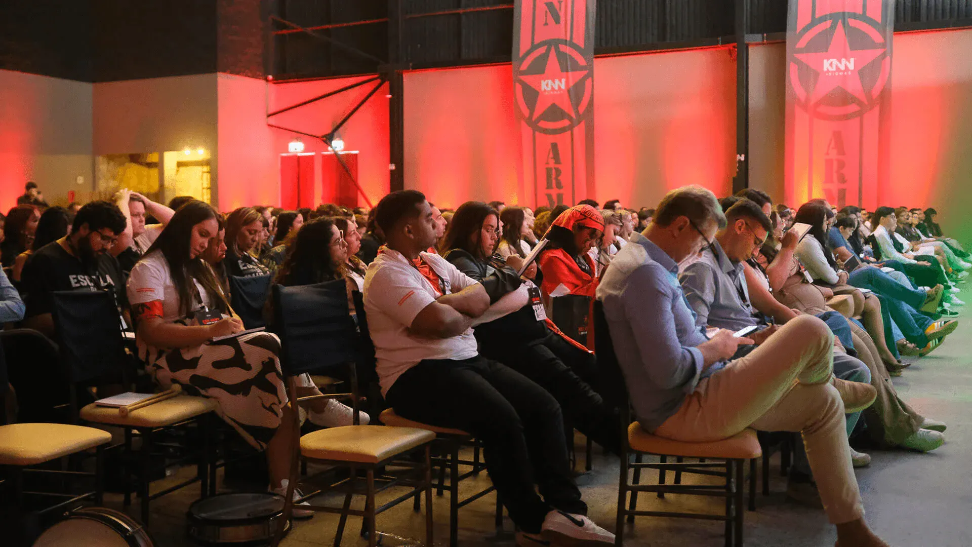 Público assistindo a evento de eSports, destaque para o ambiente com iluminação vermelha e banners com logo do KNN, incluindo jovens e adultos concentrados na apresentação.