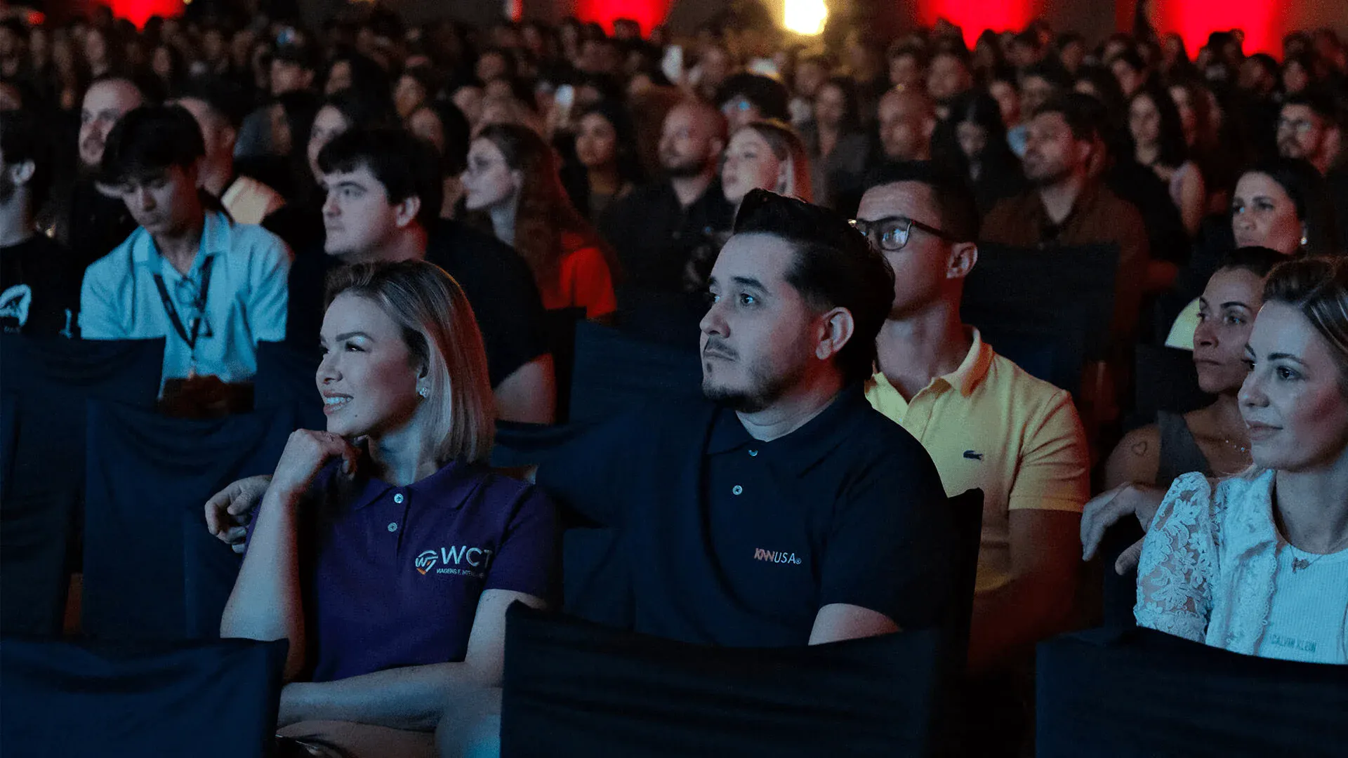Pessoa participando de evento com público interessado, sentado em cadeira no palco, com iluminação de auditório ao fundo, representando engajamento em evento corporativo ou conferência.