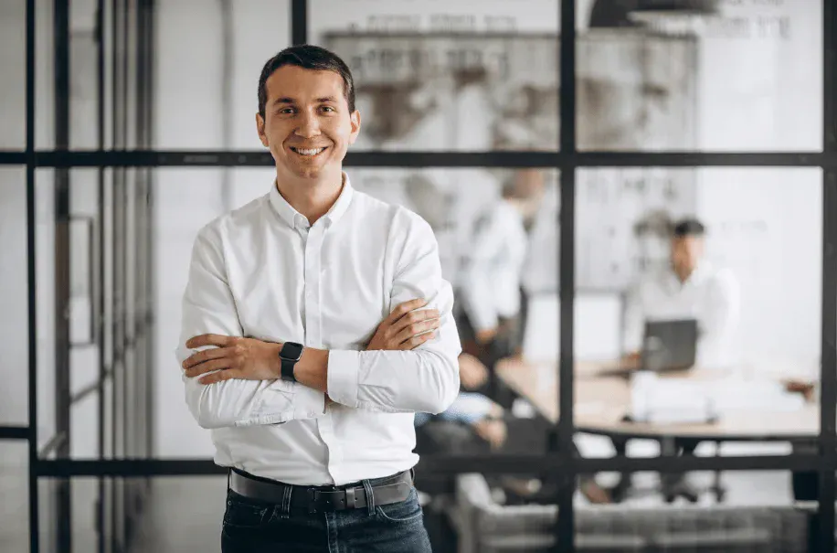 Homem sorridente posando com braços cruzados em ambiente de escritório moderno.