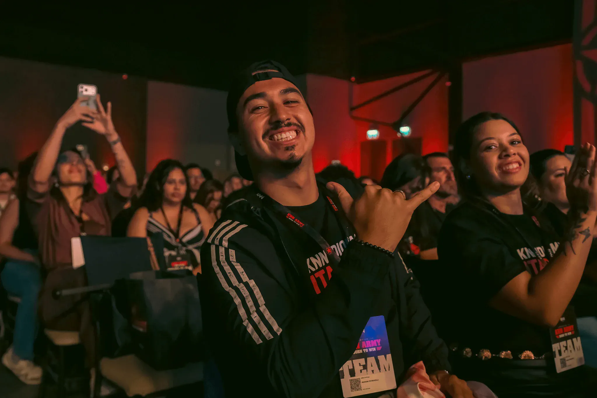 Homem sorridente em evento, rodeado de pessoas emocionadas, com iluminação de palco vermelha ao fundo, mostrando entusiasmo e alegria durante a convenção.