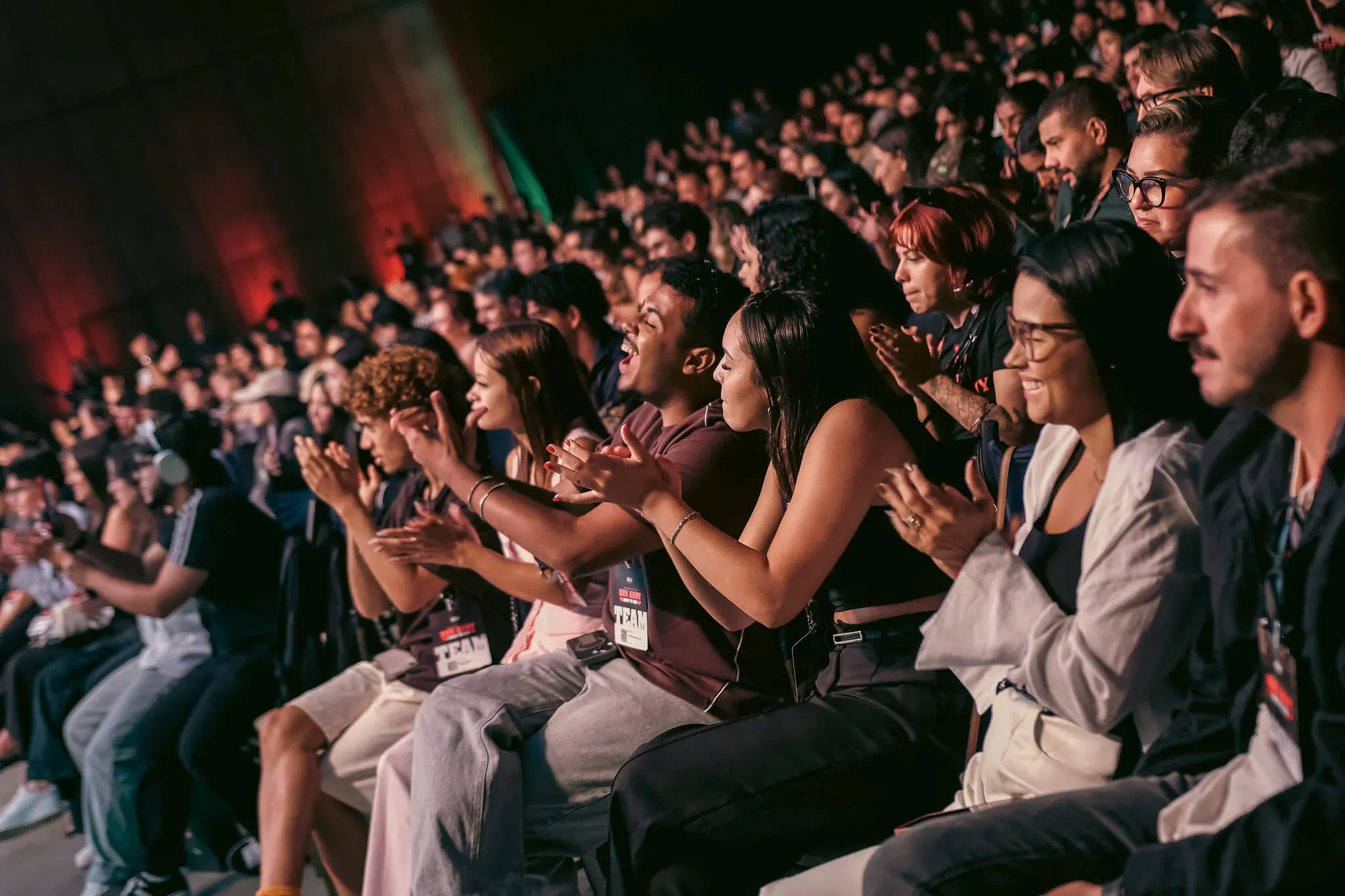 Público em um auditório lotado aplaudindo e assistindo a uma apresentação, com pessoas sentadas em fileiras; alguns usam crachá, luzes coloridas no fundo e clima de evento corporativo ou conferência.