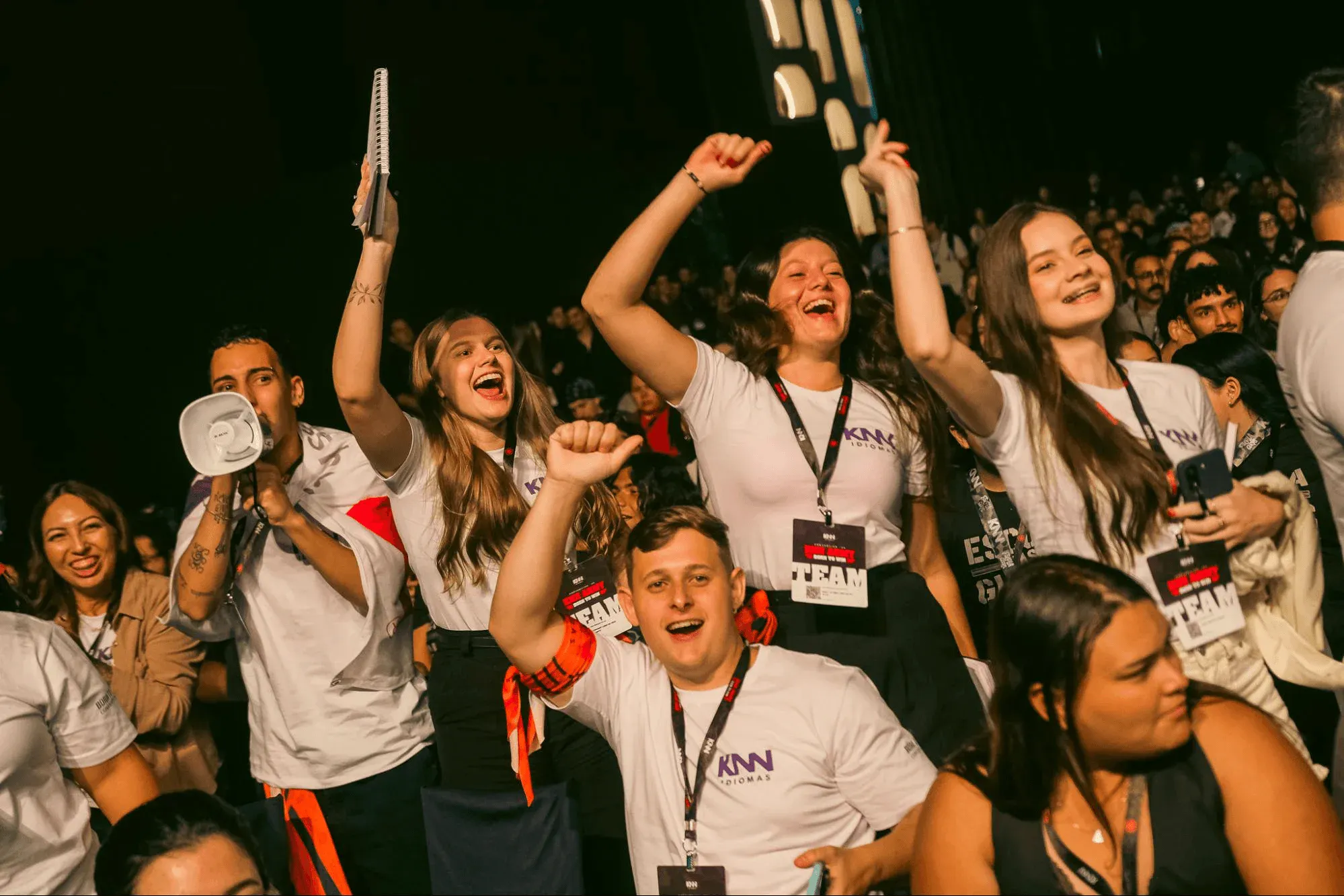 Equipe de competição de eSports comemorando em um evento ao vivo, com integrantes vestidos com camisetas do time, levantando troféus e exibindo crachá, em meio a uma plateia lotada ao fundo.