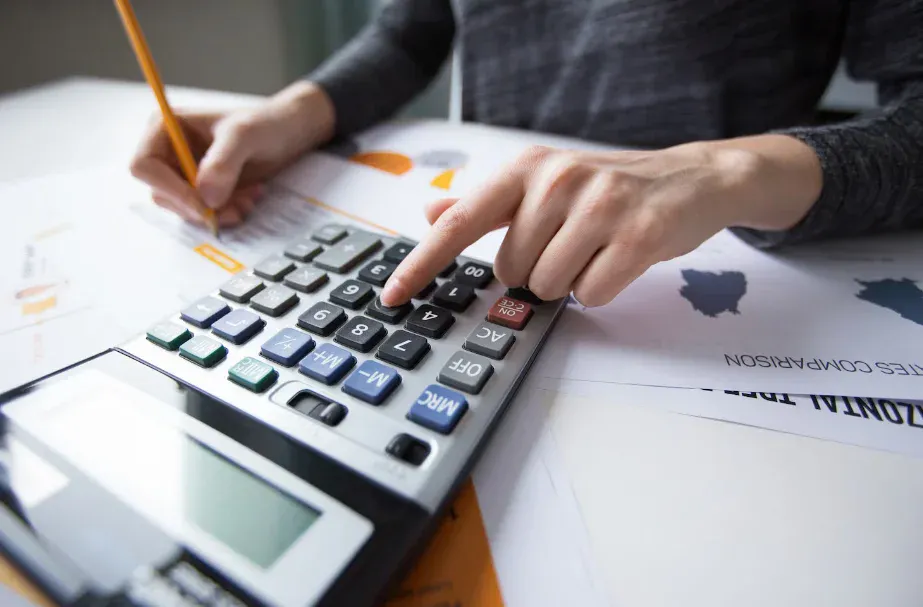 Pessoa usando uma calculadora e fazendo anotações em uma mesa de trabalho com gráficos e documentos ao fundo