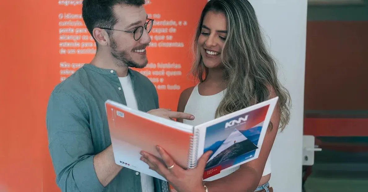 Homem e mulher sorrindo enquanto olham na apostila KNN Idiomas em uma conversa amigável