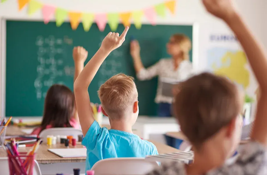 Crianças participando de aula na escola, com atenção ao professor, levantando as mãos
