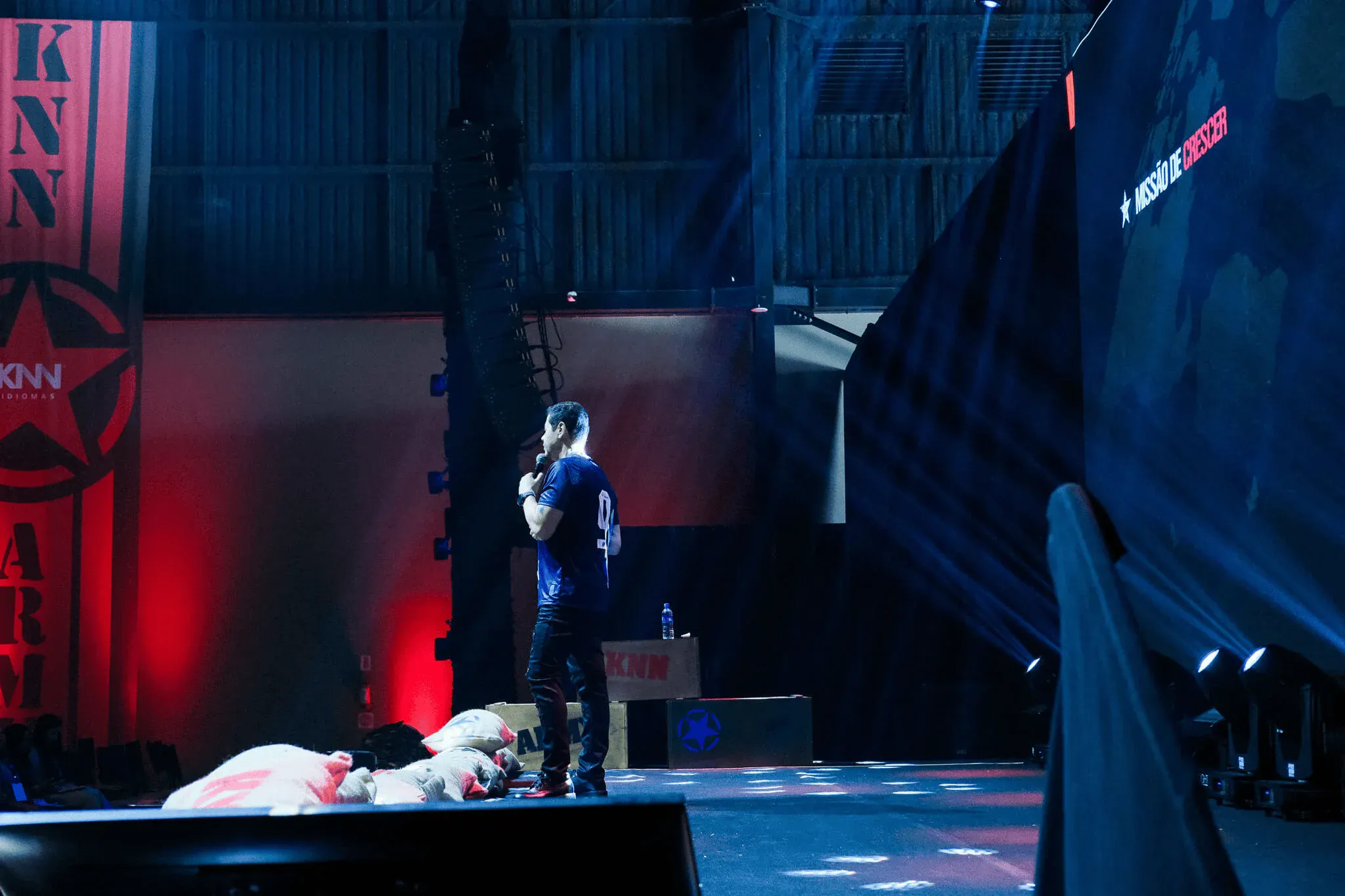 Homem em pé em um palco de evento com iluminação azul e vermelha, ao fundo aparecem estruturas e iluminação cênica e parte de uma lona de cenário, com equipamento de som ao lado.