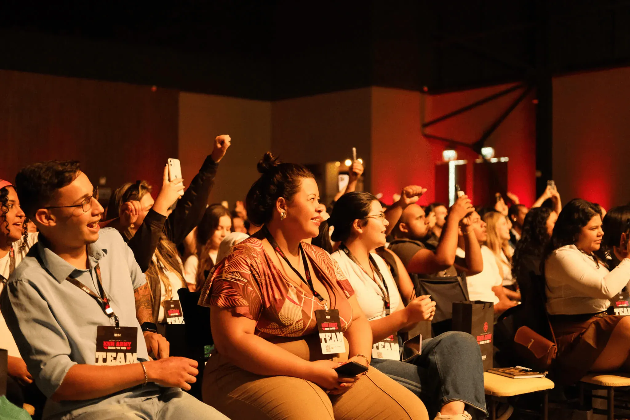 Participantes em um auditório durante um evento, com palestrantes e equipe reunidos em fileiras. Pessoas levantam a mão e acompanham palestras, usando crachás e roupas casuais.