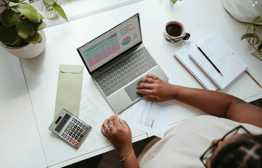 Pessoa usando um laptop com gráficos e relatórios na tela, enquanto analisa documentos financeiros sobre a mesa com calculadora, canetas e xícara de café.