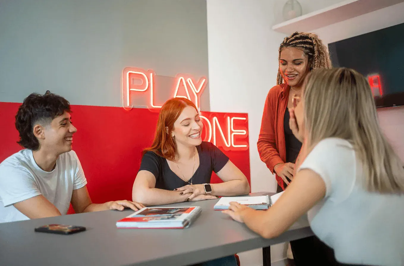 Mulher sorridente com roupa preta, segurando as mãos enquanto conversa em um ambiente interno, com luz suave e fundo desfocado.