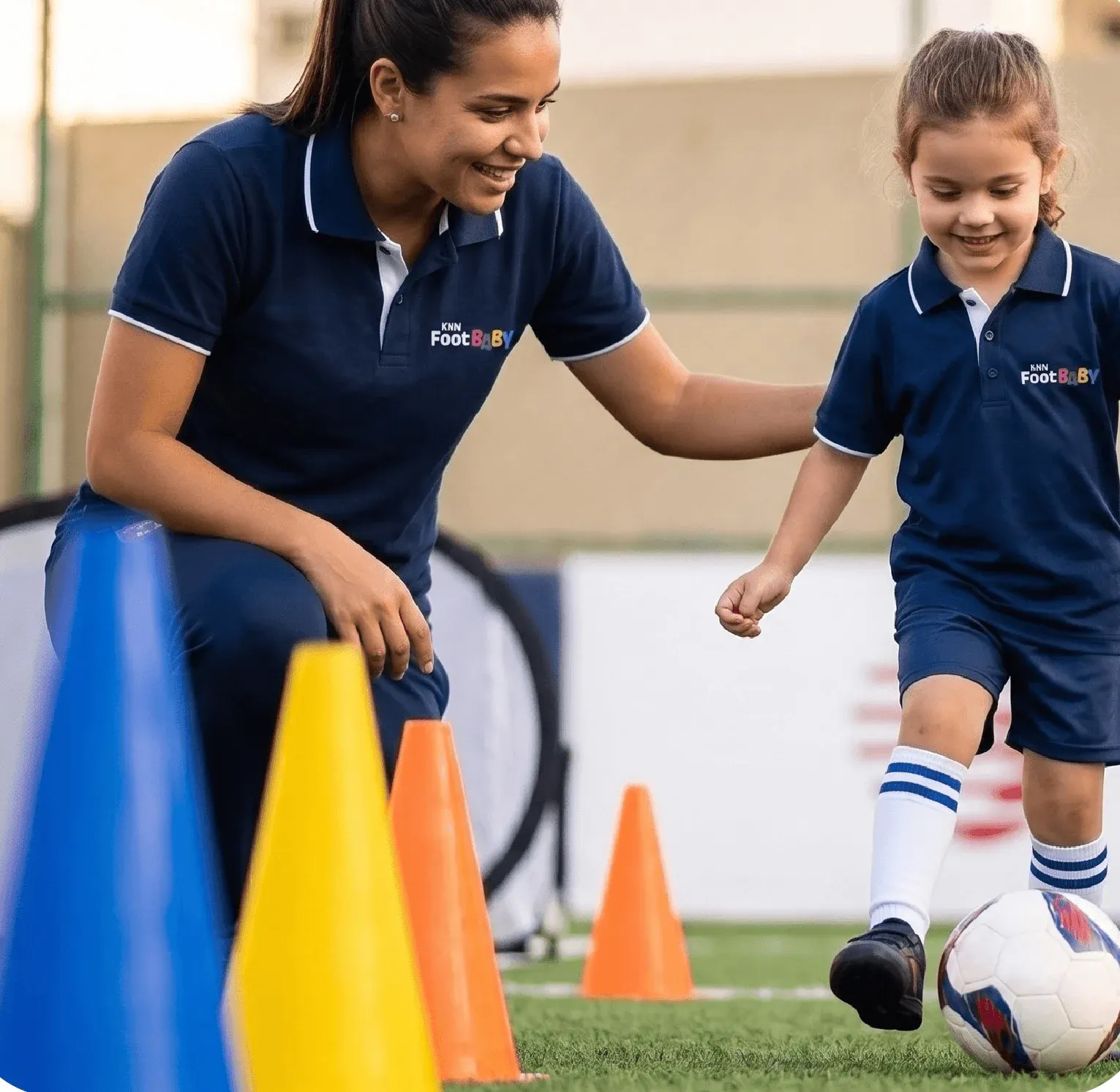 Professora e criança brincando com bola e cones em aula de futebol no gramado, usando uniformes azuis e sorrindo durante atividade esportiva.