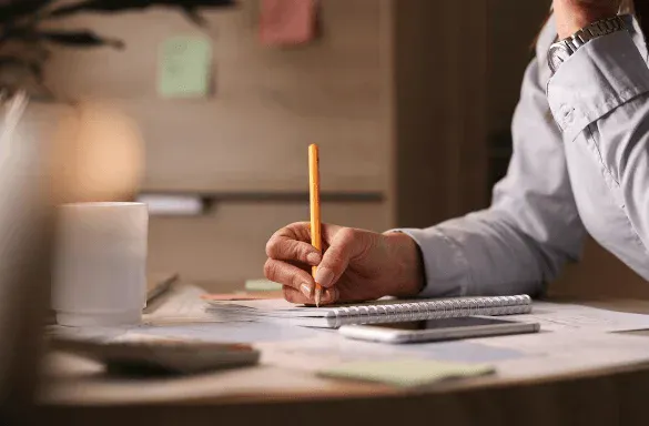 Pessoa assinando um documento com caneta amarela em uma mesa de escritório, com papéis, caderno e itens de trabalho ao fundo.