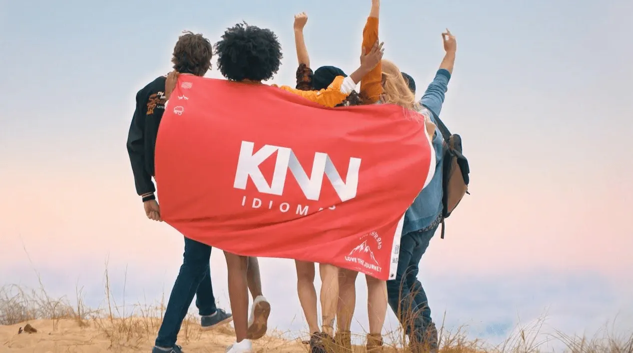 Grupo de pessoas em uma trilha no deserto com bandeira vermelha “KNW IDIOM” nas costas, braços erguidos e pôr do sol ao fundo.