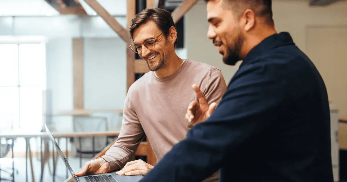 Dois homens trabalhando em uma cozinha compartilhada, um deles sorrindo enquanto usa um notebook, transmitindo uma atmosfera de colaboração e rotina profissional.